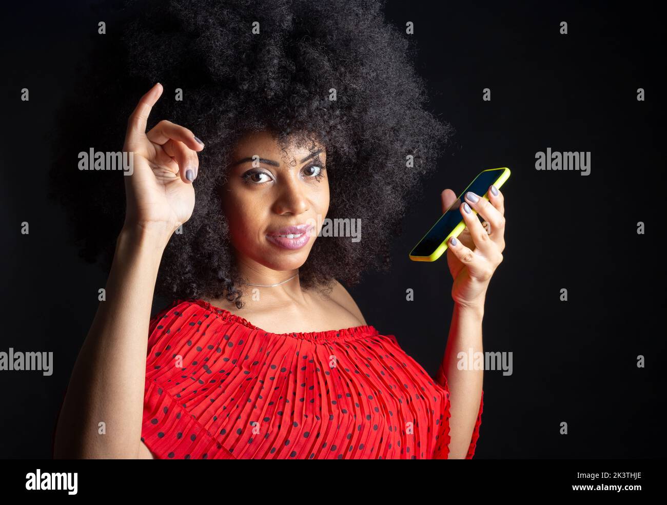 Giovane ragazza etnica allegra con cellulare e acconciatura afro con mano rialzata mentre si guarda la macchina fotografica Foto Stock
