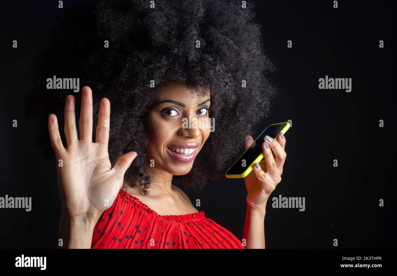 Giovane ragazza etnica allegra con cellulare e acconciatura afro dicendo ciao con mano sollevata mentre si guarda la macchina fotografica Foto Stock