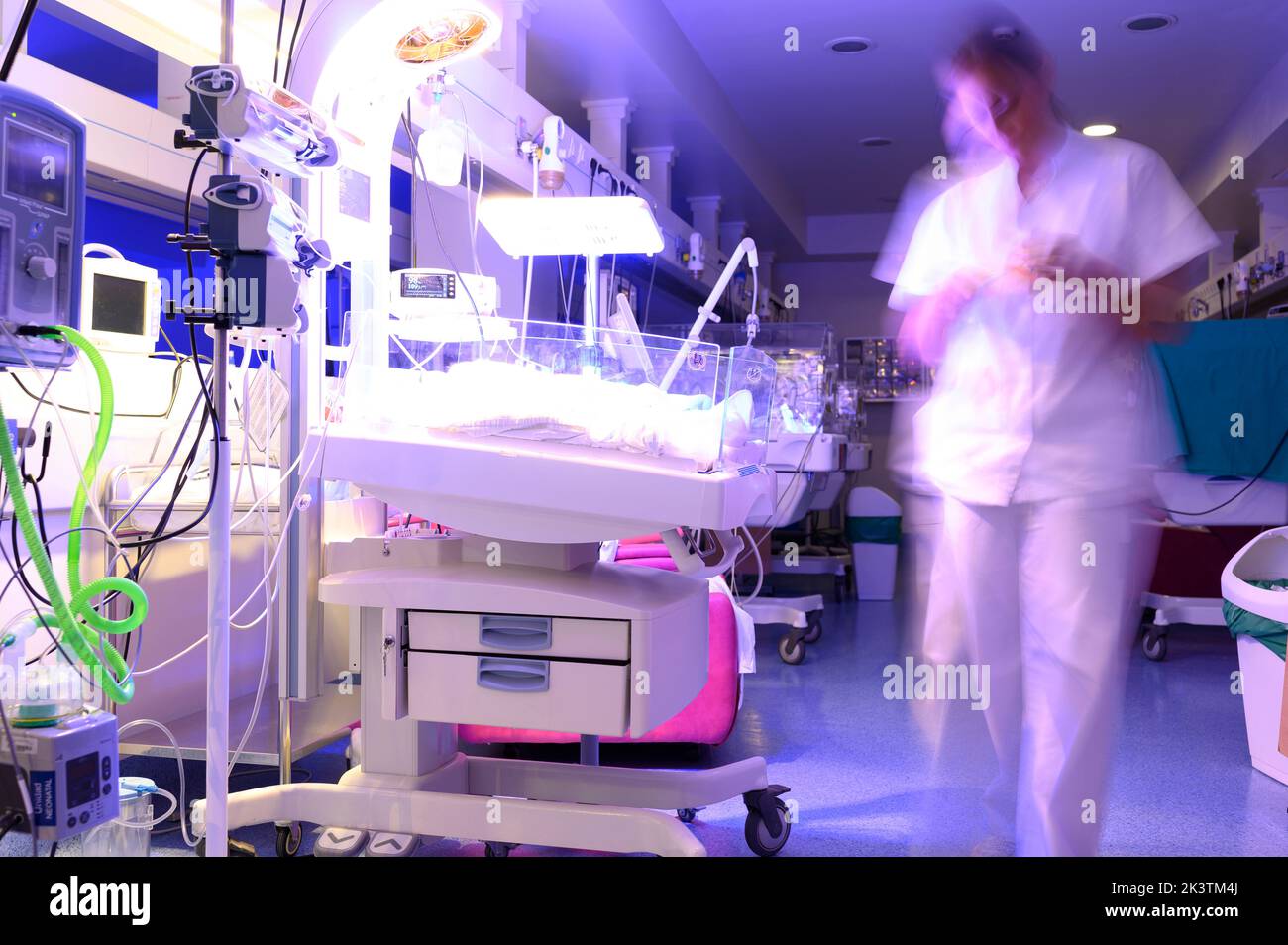 Donna sfocata in uniforme medica bianca camminando vicino ad incubatori durante il lavoro in ospedale neonatale di notte Foto Stock