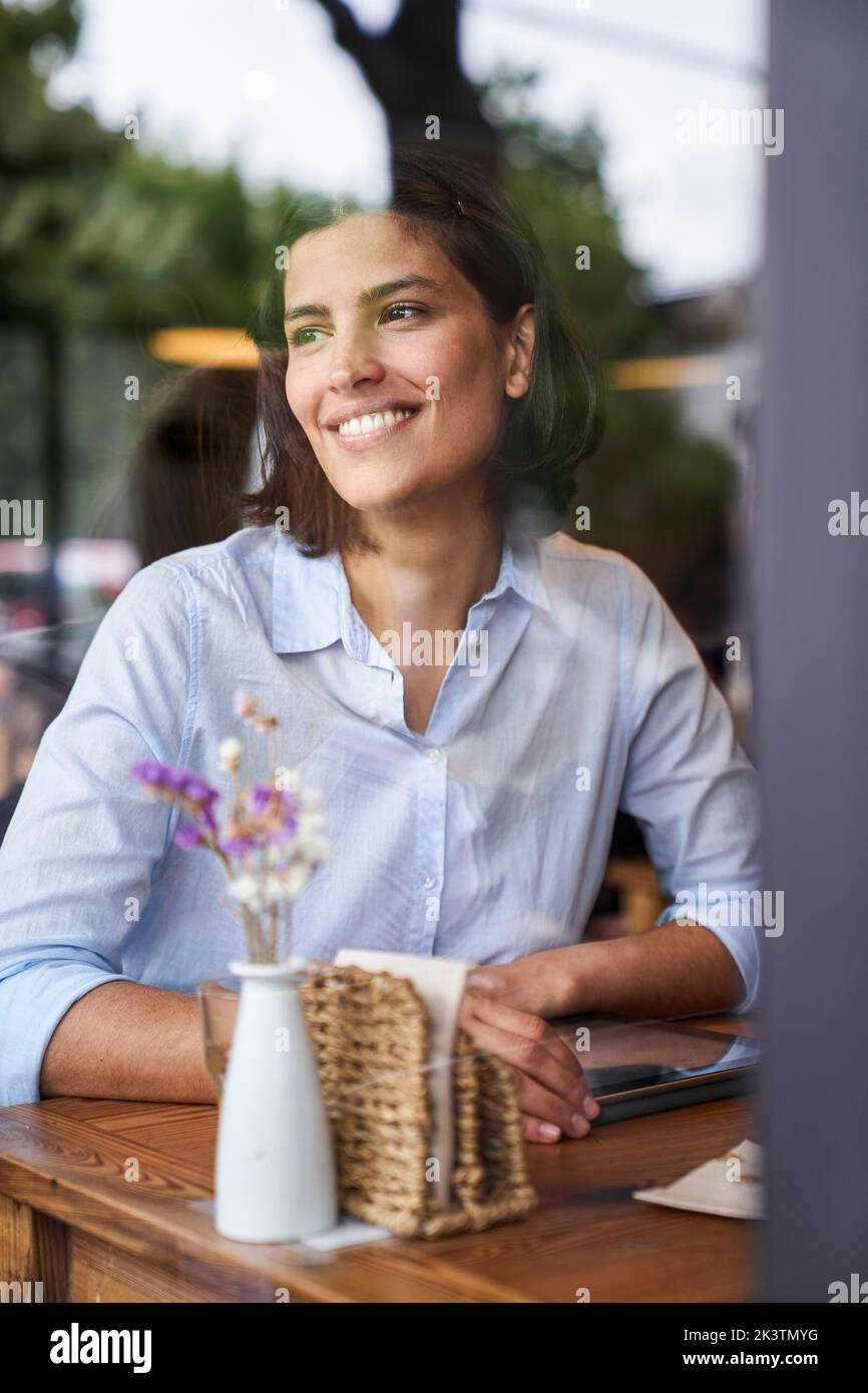Ritratto girato attraverso la finestra di una donna sorridente latino-americana seduta al caffè Foto Stock