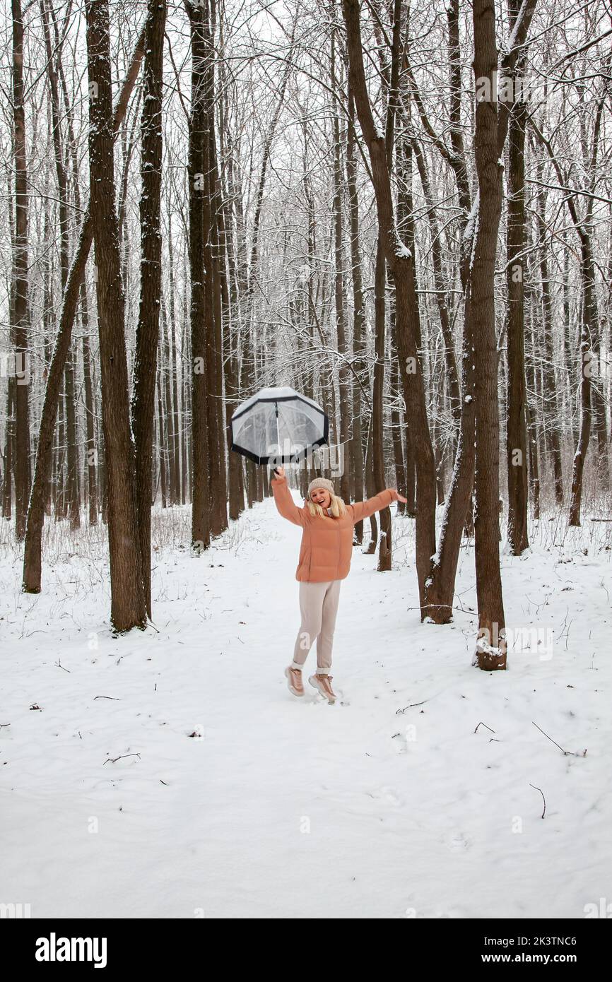Una giovane donna salta in una pineta con un ombrello trasparente, cercando di decolorare, gioendo in inverno. Foto Stock
