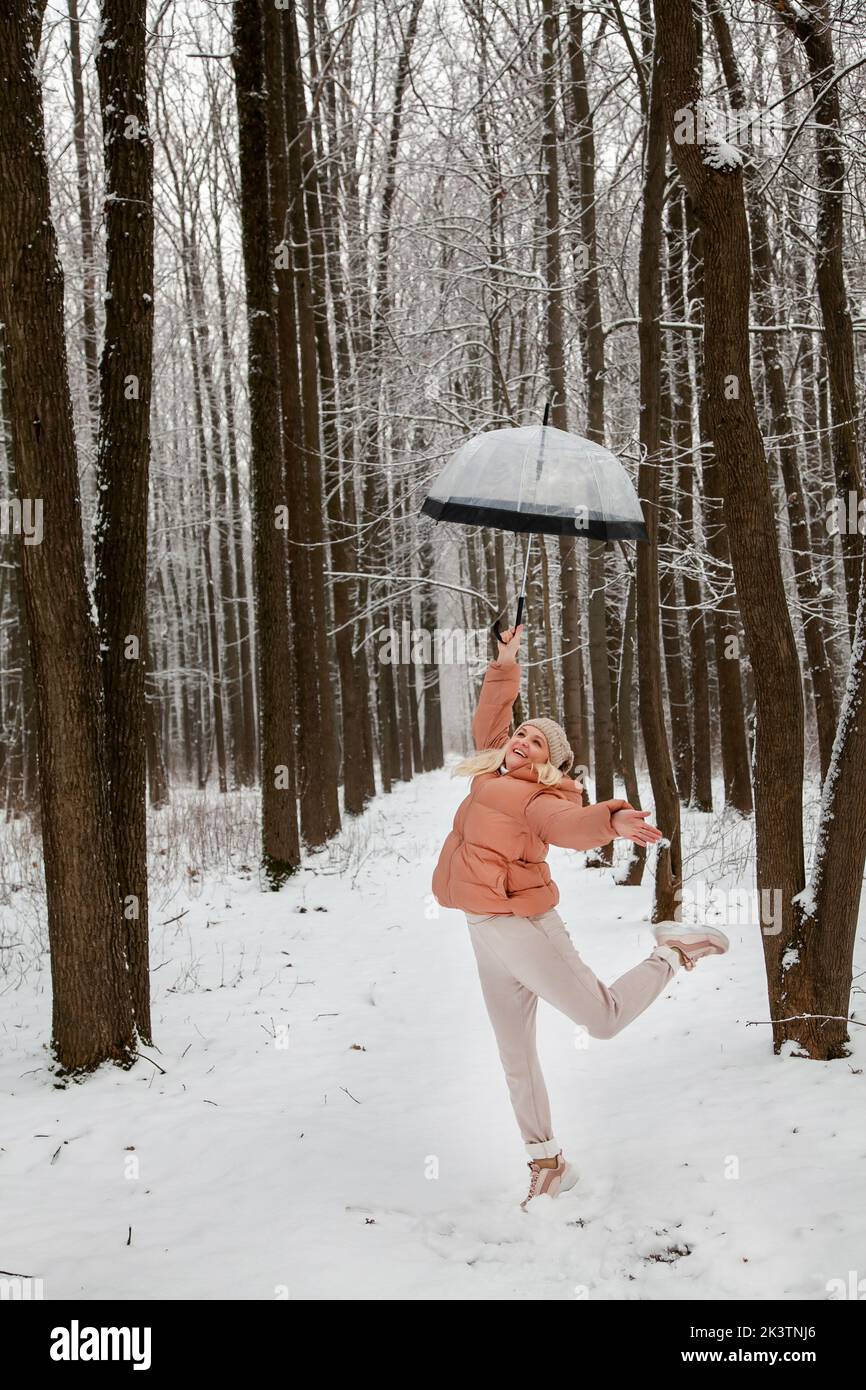 Una giovane donna salta in una pineta con un ombrello trasparente, cercando di decolorare, gioendo in inverno. Foto Stock
