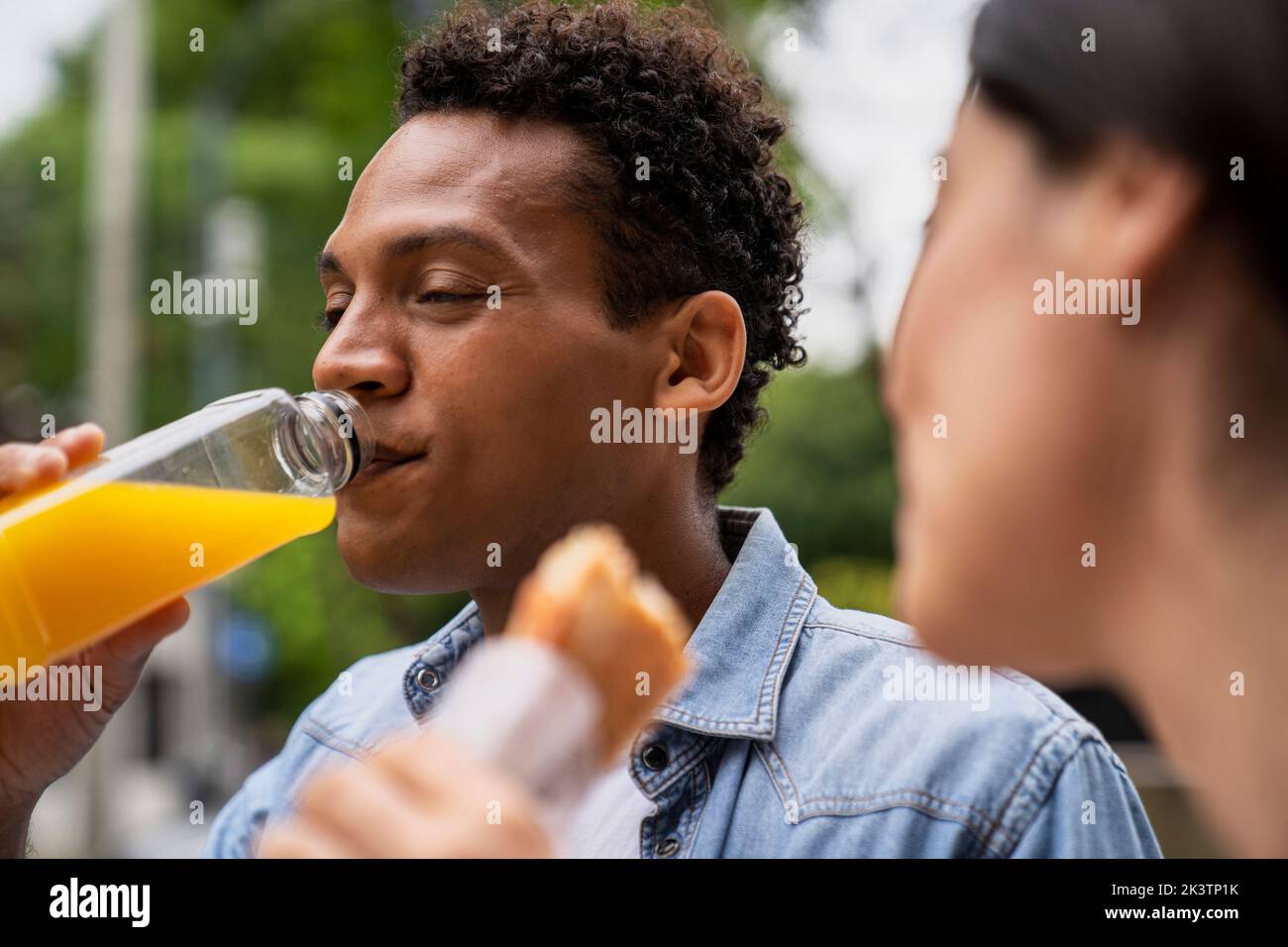 Vista laterale di un uomo afroamericano che beve succo d'arancia e la sua amica latinoamericana che mangia un panino Foto Stock
