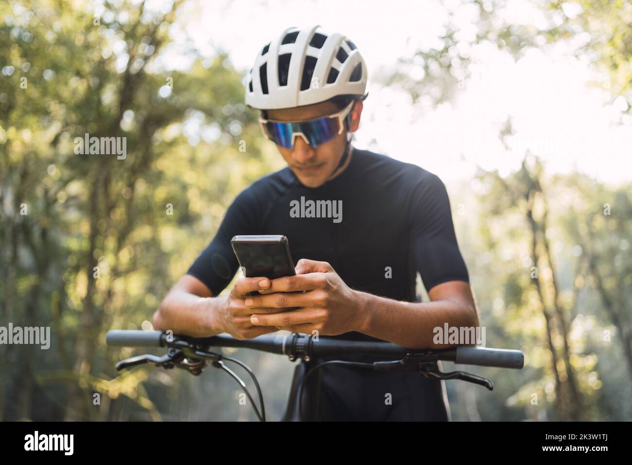 Biker maschile in casco e occhiali da bicicletta messaggi di testo sul cellulare mentre si siede in bicicletta nel bosco nelle giornate di sole Foto Stock