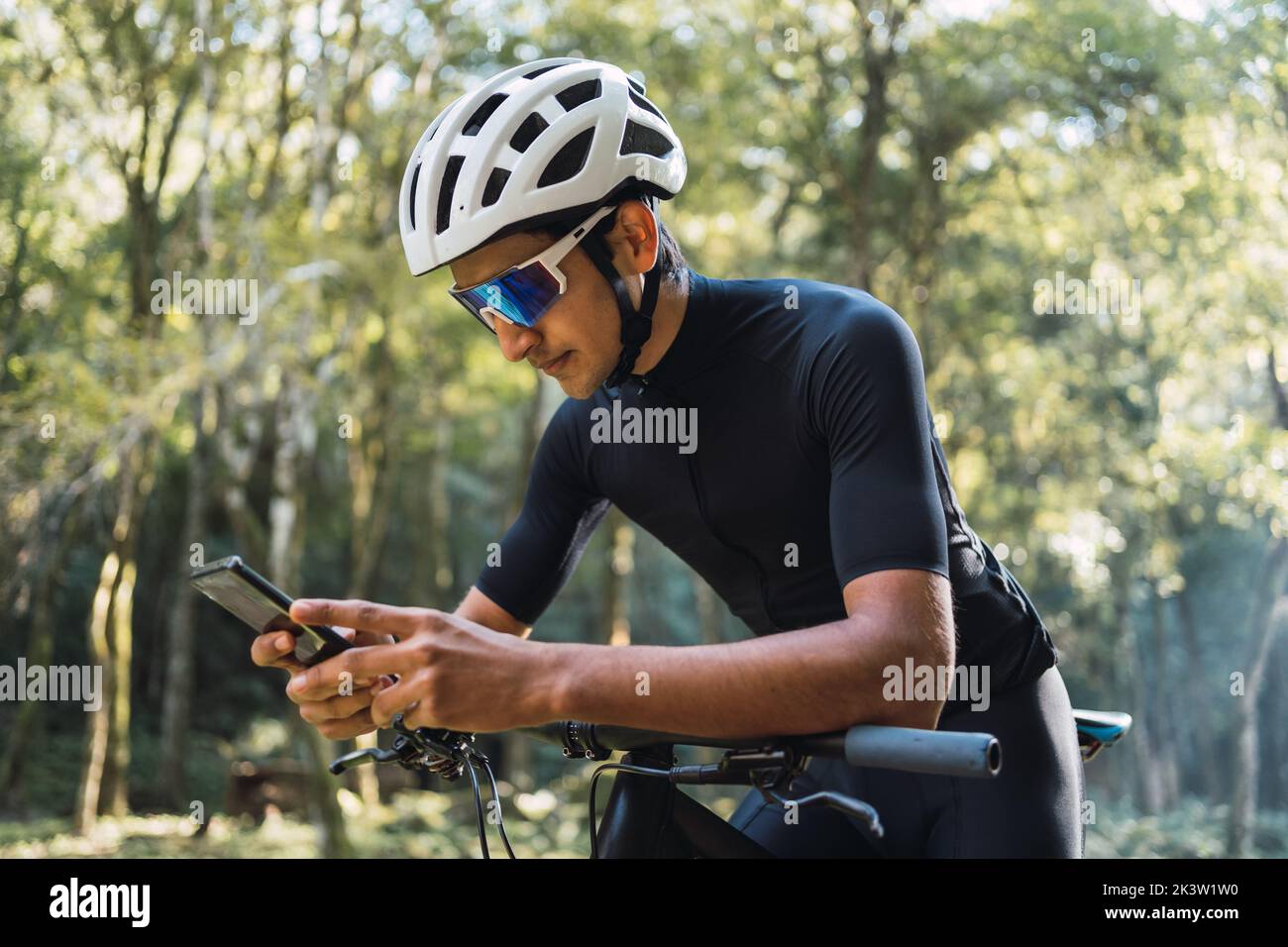 Biker maschile in casco e occhiali da bicicletta messaggi di testo sul cellulare mentre si siede in bicicletta nel bosco nelle giornate di sole Foto Stock