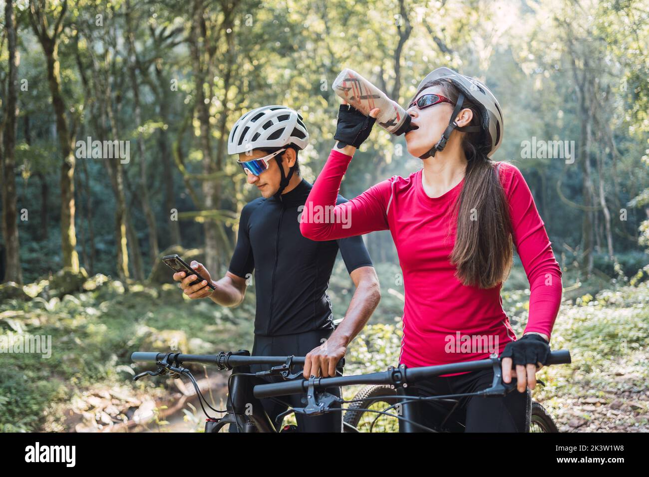 Ciclista femmina acqua potabile contro amico maschio surfing internet sul cellulare durante il viaggio in bicicletta nel bosco Foto Stock