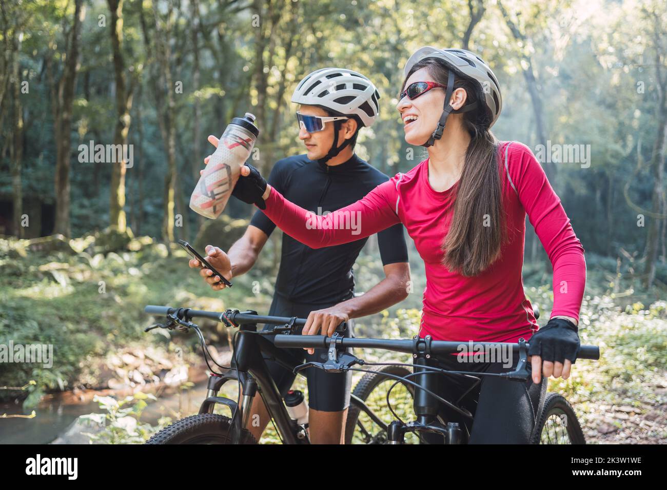 Ciclista femmina acqua potabile contro amico maschio surfing internet sul cellulare durante il viaggio in bicicletta nel bosco Foto Stock