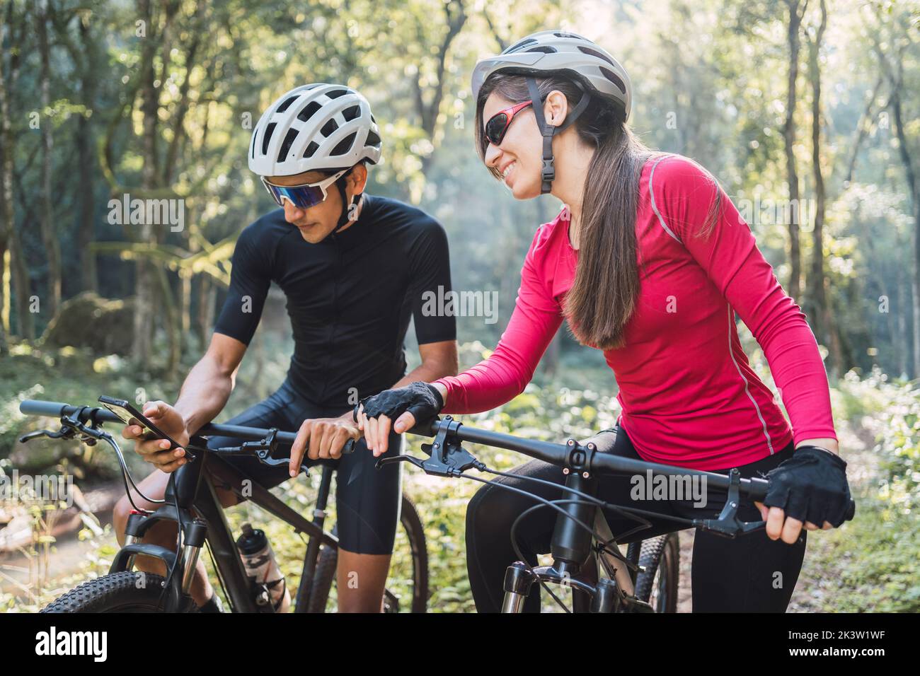 Sorridente sportswoman in occhiali da bicicletta parlando con un amico maschio con il cellulare controllare la direzione con GPS app in bicicletta durante il viaggio nella foresta Foto Stock