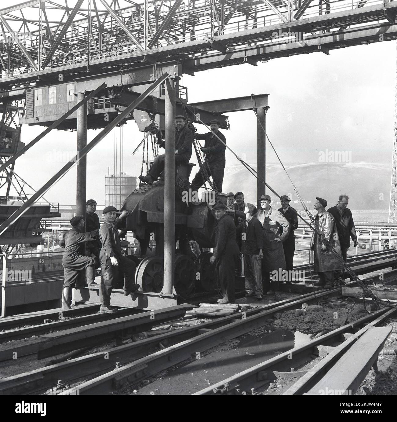 Anni '1950, storico, un gruppo di operai siderurgici riuniti da un argano su un binario di manovra su rotaia, sotto un cavalletto con struttura in acciaio, Port Talbot, Galles, Regno Unito. Foto Stock