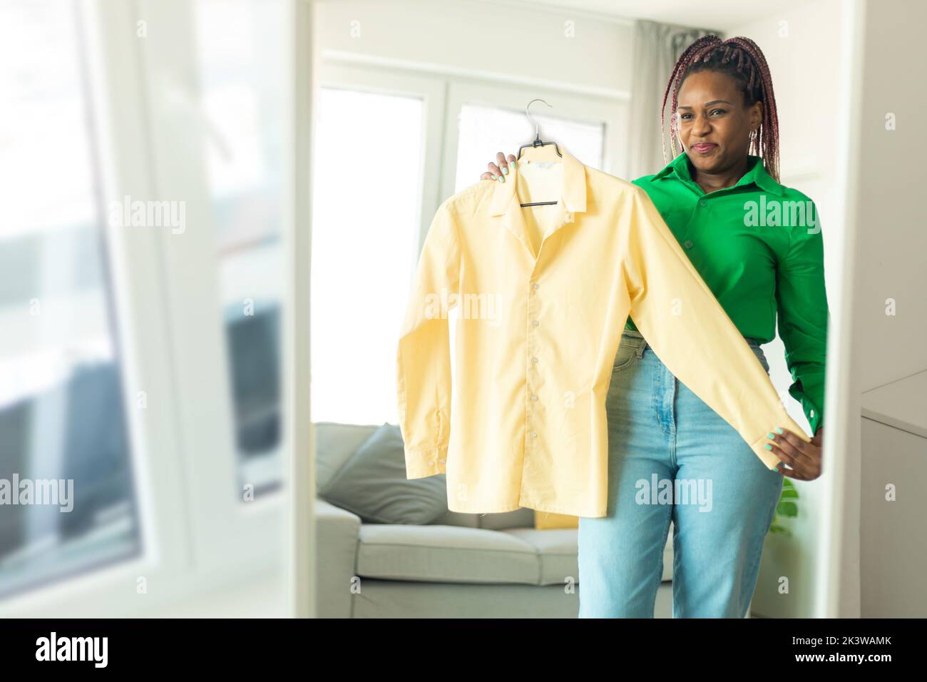 Donna afroamericana scegliendo i vestiti da indossare, guardando se stessa nello specchio in soggiorno a casa, spazio libero Foto Stock