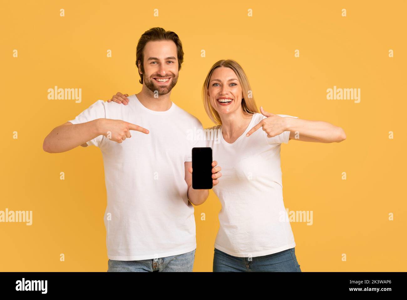 Felice giovane coppia caucasica in t-shirt bianche punta le dita sullo smartphone con schermo vuoto e consiglia app Foto Stock