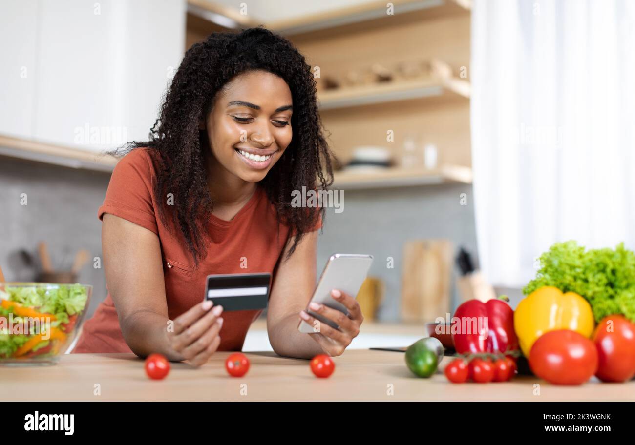 Allegro bella millennial nero femmina con smartphone e carta di credito godere di acquistare ordine a tavolo Foto Stock