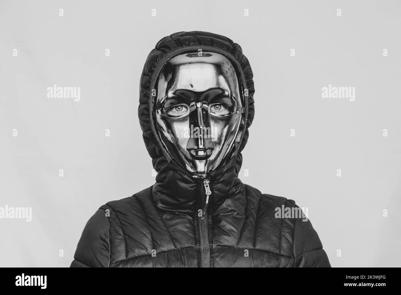 Ragazza in una maschera di plastica senza volto e giacche con un cappuccio su uno sfondo bianco foto in bianco e nero, anonimato, mistero Foto Stock