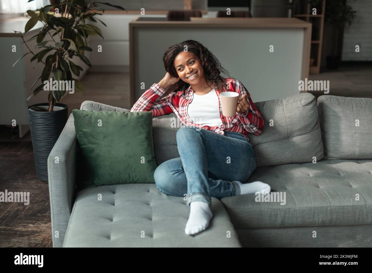 Allegra ragazza afro-americana millenaria con una tazza di bevanda preferita, goditi il tempo libero e il comfort, siediti sul divano Foto Stock