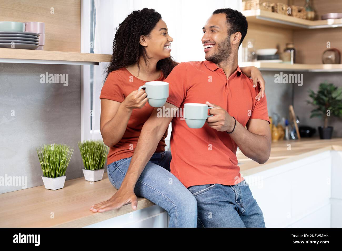 Sorridente donna nera millenaria che abbraccia l'uomo, allietano le tazze con il caffè, godono di tempo libero in cucina interna Foto Stock