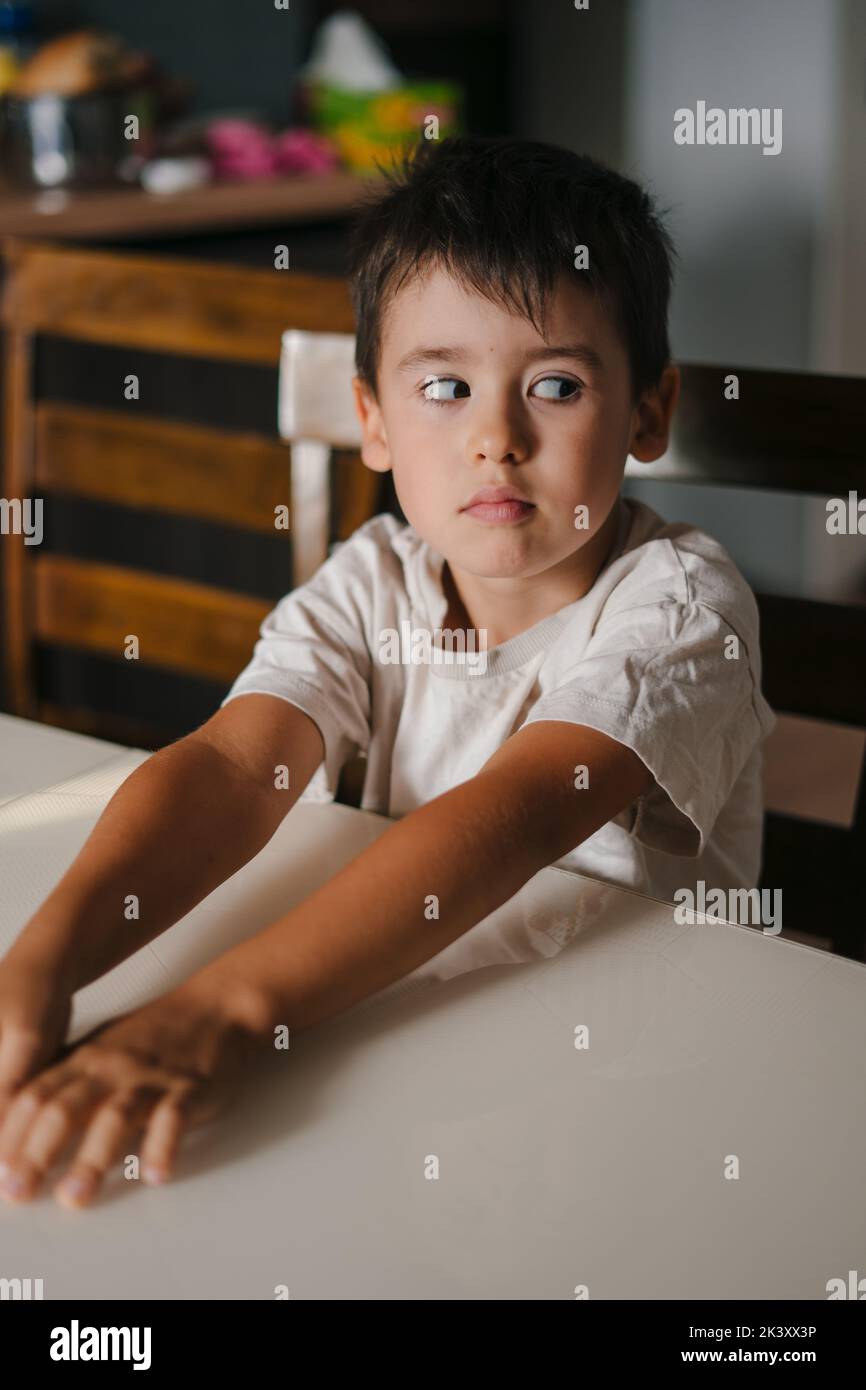 Ragazzo allegro seduto al tavolo in cucina e aspetta che sua madre gli faccia colazione la mattina. La famiglia trascorre del tempo insieme a casa nel fine settimana Foto Stock