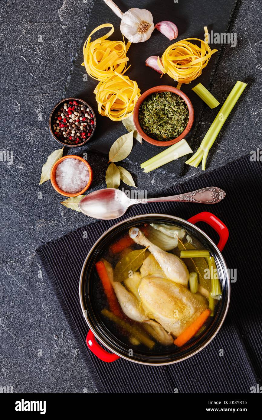 brodo di pollo cotto con pollo intero, sedano, cipolla e carota in pentola rossa con ingredienti e tagliatelle su tavolo di cemento, vista verticale Foto Stock