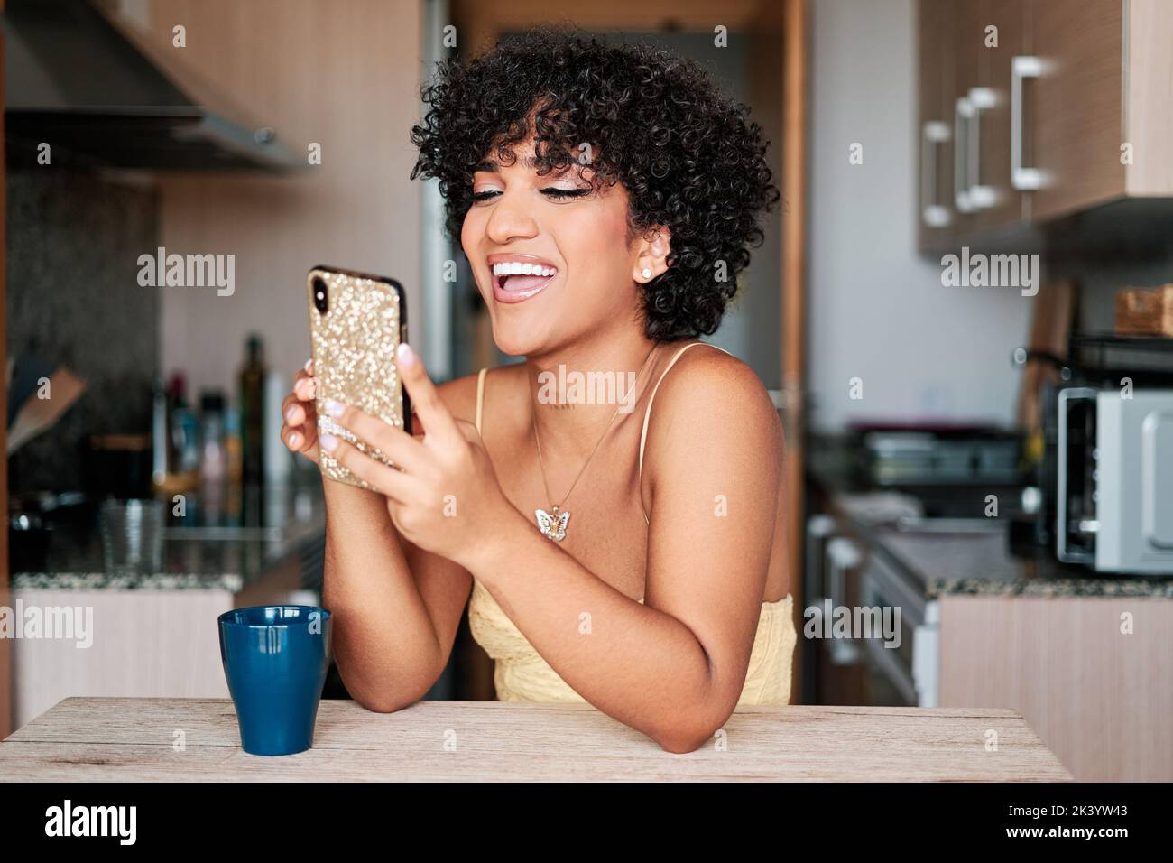 Donna transgender felice ridendo mentre usando un telefono mobile seduto al tavolo della cucina a casa. Foto Stock