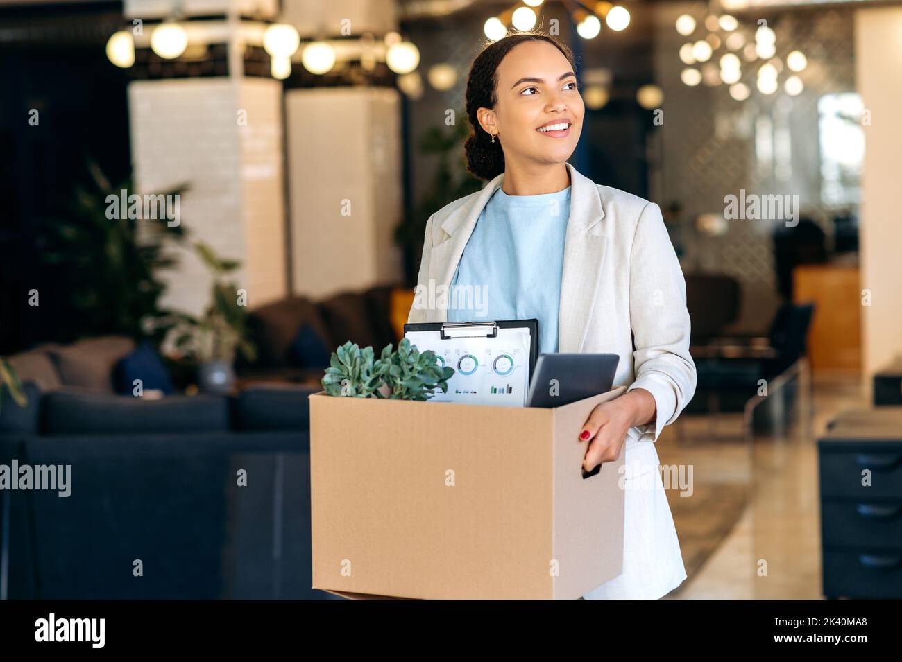 Nuovo lavoro. Eccitata motivata di successo gara mista donna, entra in un nuovo ufficio di lavoro, tenendo una scatola di cartone con attributi di lavoro nelle sue mani, ispeziona lo spazio di lavoro con stupore, sorridendo felicemente Foto Stock