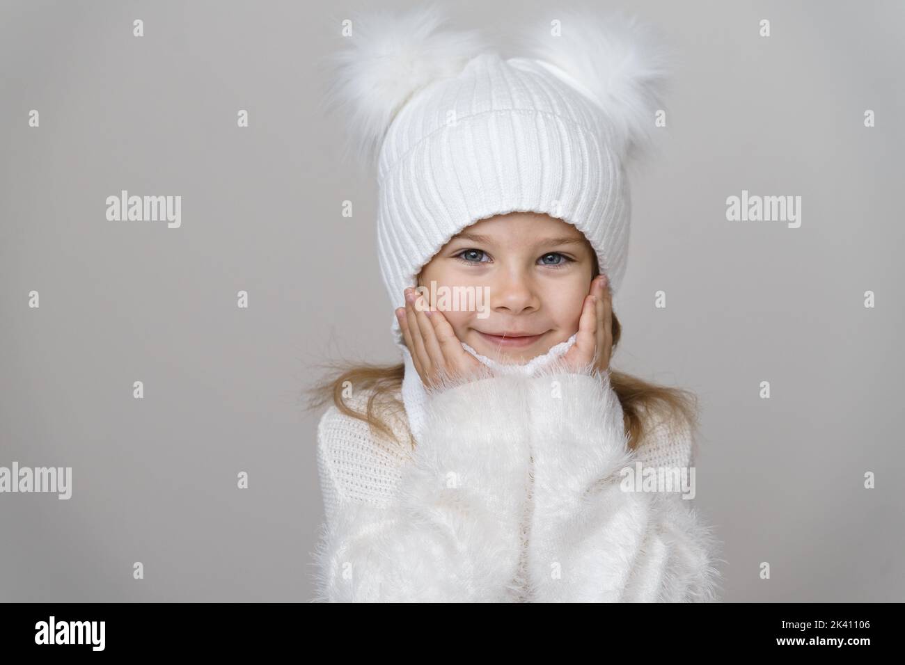 Ragazza carina di 6 anni in un cappello di lana bianco, maglione e snood su sfondo grigio. Abiti caldi invernali. Foto Stock