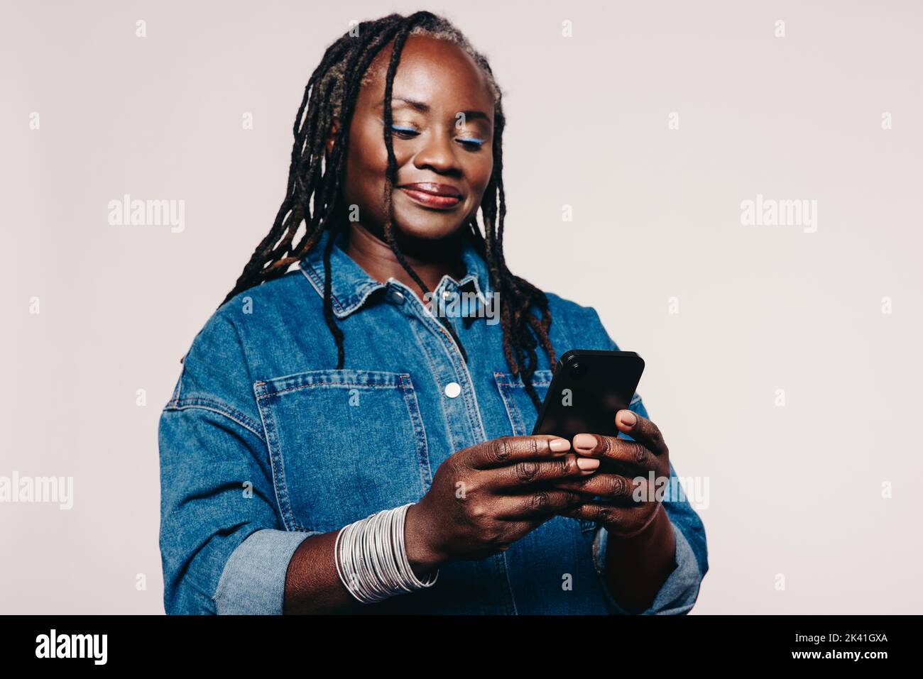Donna matura che legge un messaggio di testo su uno smartphone mentre si trova su uno sfondo grigio. Donna elegante con lucchetti che indossano una giacca in denim e. Foto Stock