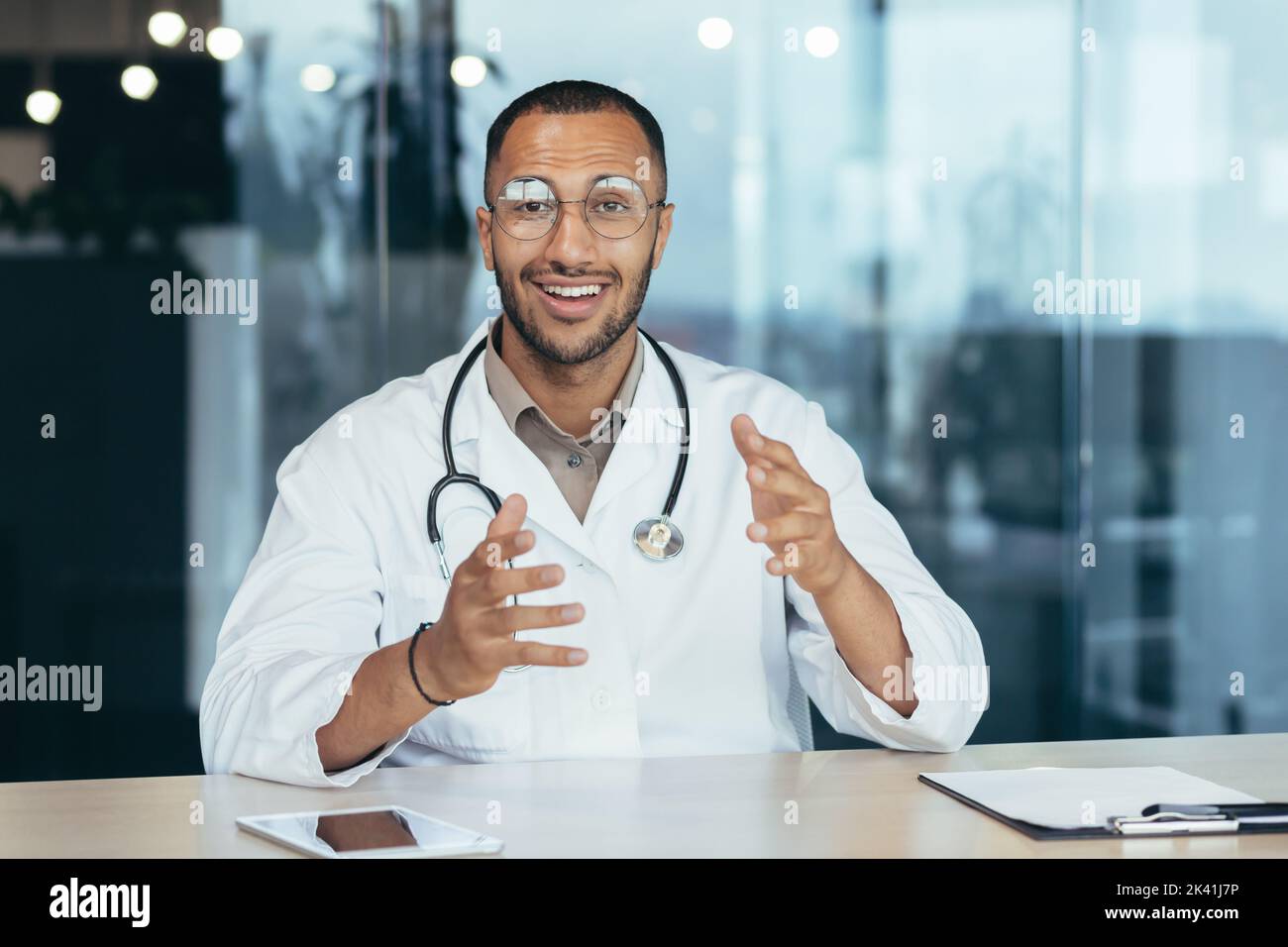 Webcam vista, medico di famiglia allegro e di successo consulta i pazienti in remoto online, videochiamata di un uomo in cappotto medico sorridente e guardando la fotocamera, lavorando all'interno di clinica ufficio. Foto Stock