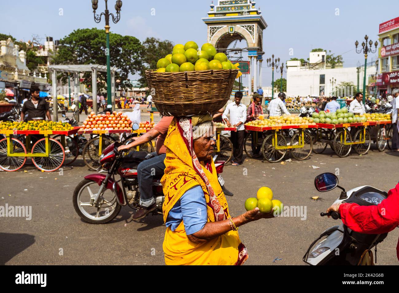 Mysore, Karnataka, India : Una donna anziana porta un cesto di frutta sulla sua testa accanto alla Dufferin Clock Tower fuori del mercato Devaraja. Foto Stock