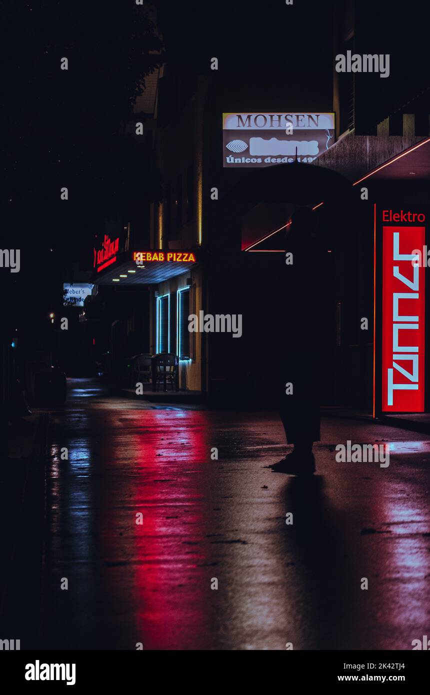 Vita notturna al neon per le strade. Segnali luminosi al neon che si riflettono sulla strada bagnata/piovosa. Insegne al neon che creano la silhouette di una persona. Foto Stock