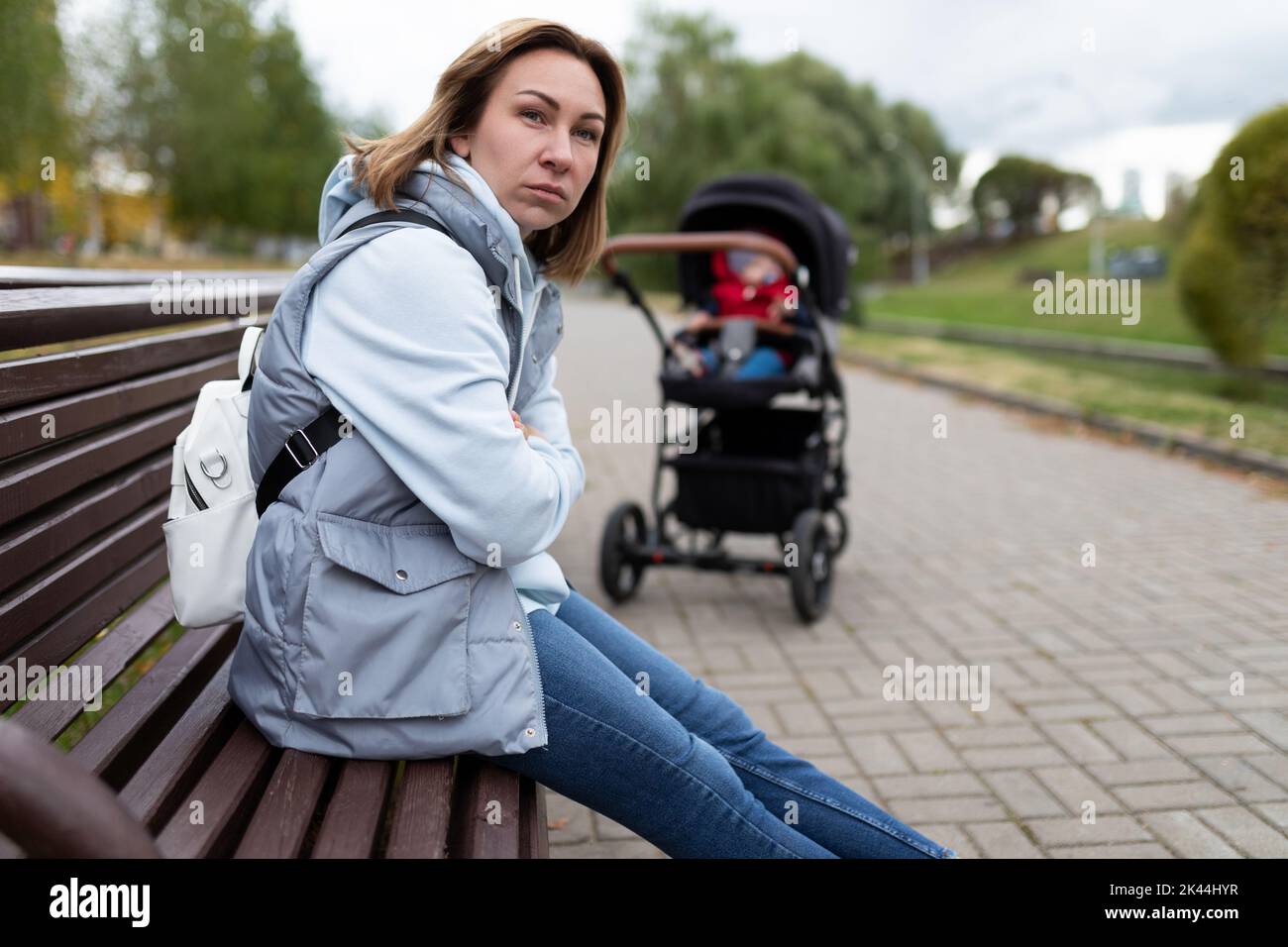 giovane madre infastidita seduta nel parco su una panchina lontano dal passeggino con un viso arrabbiato. il concetto di depressione postpartum Foto Stock