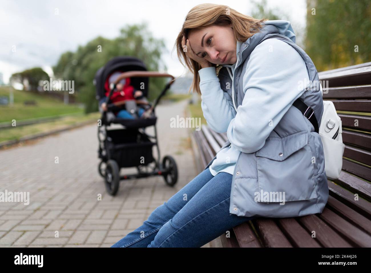 giovane donna madre di depressione postpartum siede con sentimenti incorporati nel parco su una panchina accanto a un passeggino Foto Stock