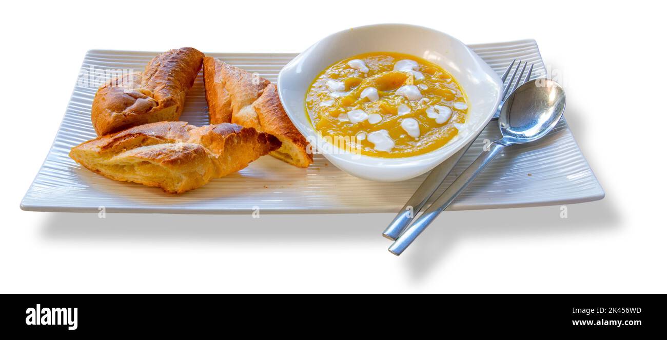 Su un piatto bianco allungato si trova una piccola ciotola di zuppa di zucca con argenteria e pane al forno. Foto Stock