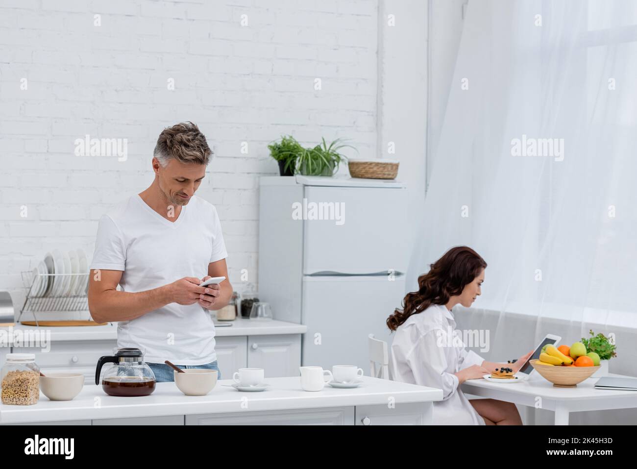 Coppia utilizzando dispositivi vicino a colazione e caffè in cucina, immagine stock Foto Stock