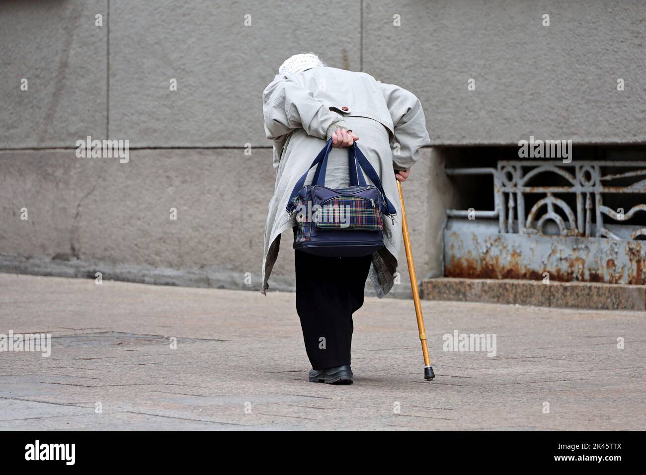 Donna anziana che cammina piegata in basso con una canna da zucchero e una borsa su una strada cittadina. Vita degli anziani in autunno, malattie della spina dorsale Foto Stock