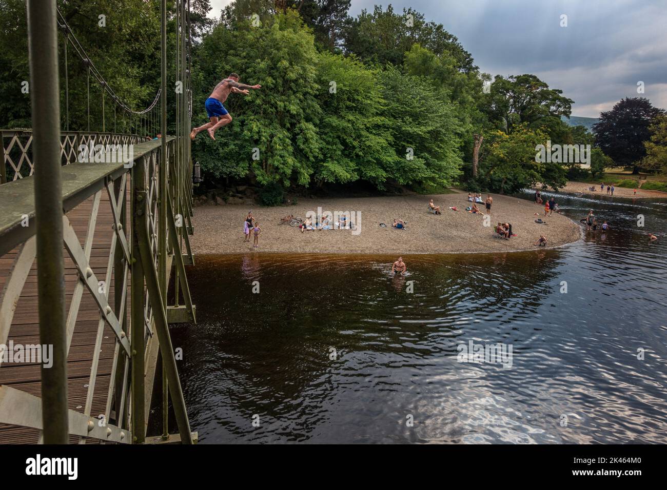 In una giornata calda sul fiume Wharfe a Ilkley, potrai goderti la spiaggia di ciottoli, con una persona che si scoppia (saltando) dal ponte sospeso. West Yorksh Foto Stock