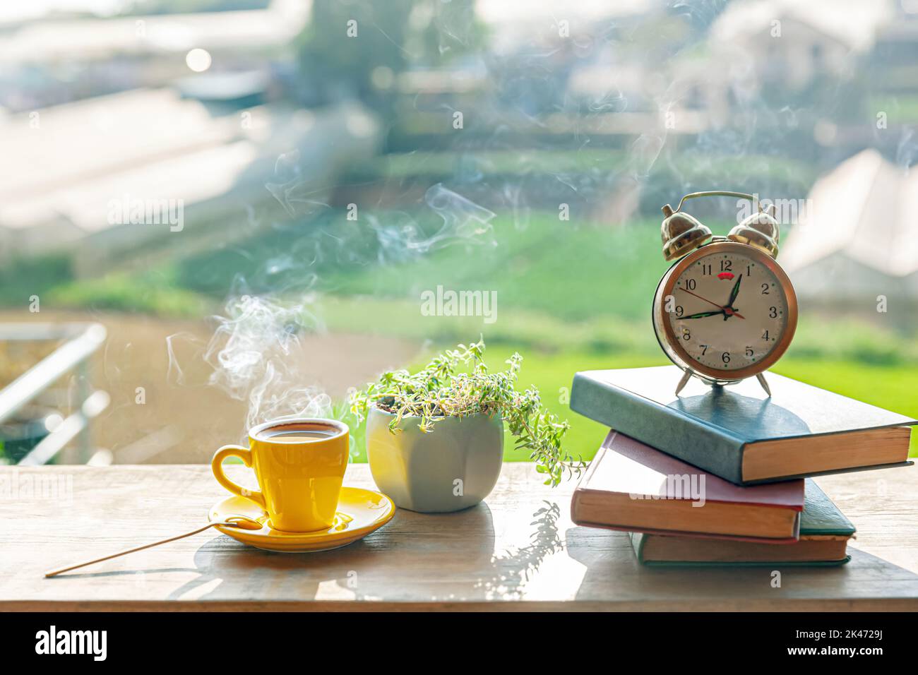 Una tazza di caffè caldo si trova accanto a una pianta in vaso, libri, sveglia su un tavolo di legno, al sole del pomeriggio Foto Stock