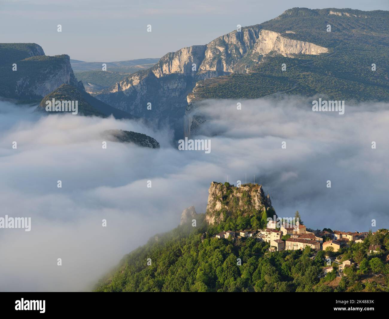 Borgo medievale arroccato sopra la nebbia mattutina con le massicce scogliere della Gola del Verdon per lo sfondo. Rougon, Alpes de Haute-Provence, Francia. Foto Stock