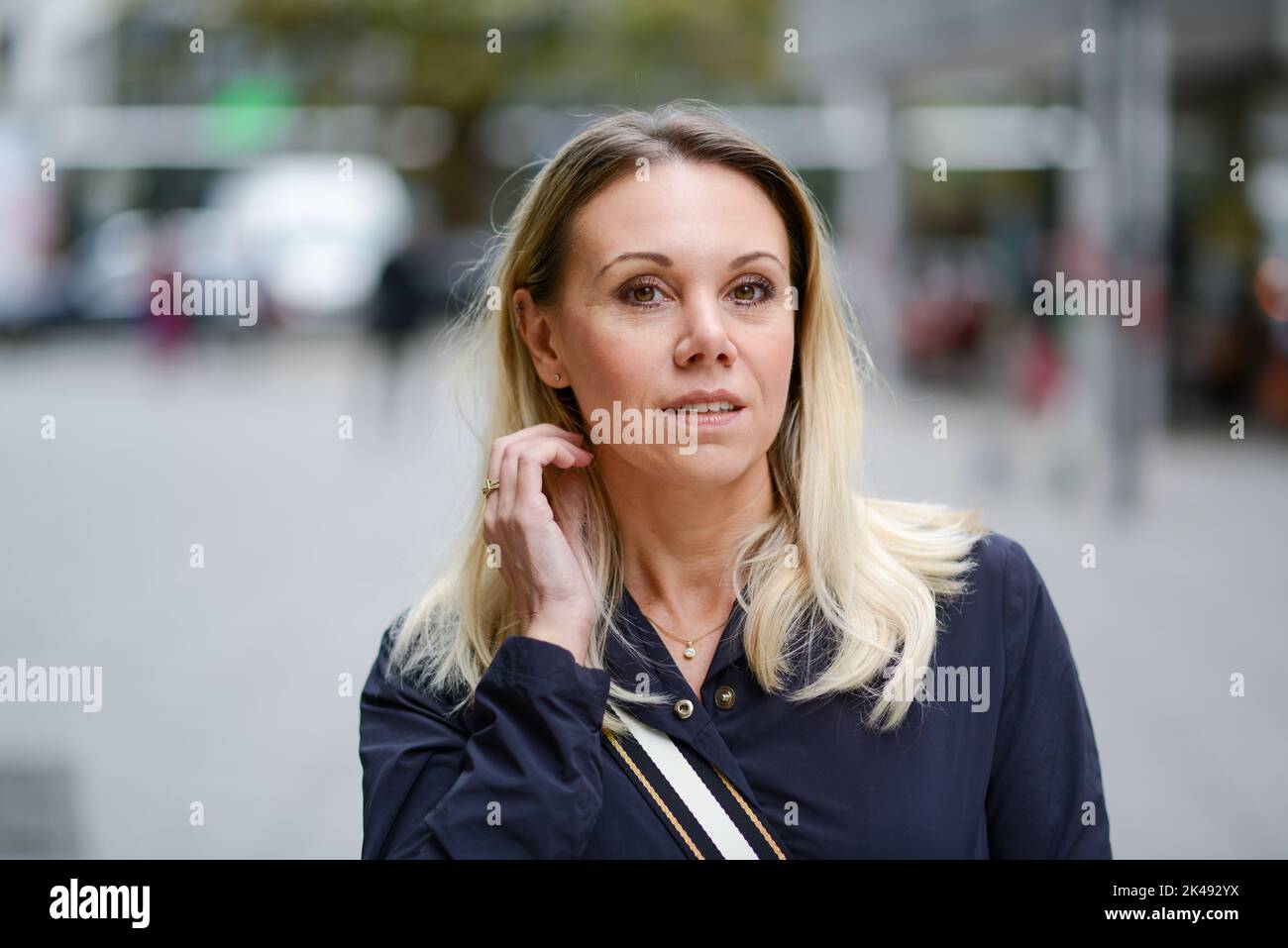 Bella donna bionda di mezza età che indossa una giacca blu guarda la macchina fotografica con un sorriso leggero mentre corre una mano attraverso i suoi capelli durante lo shopping Foto Stock