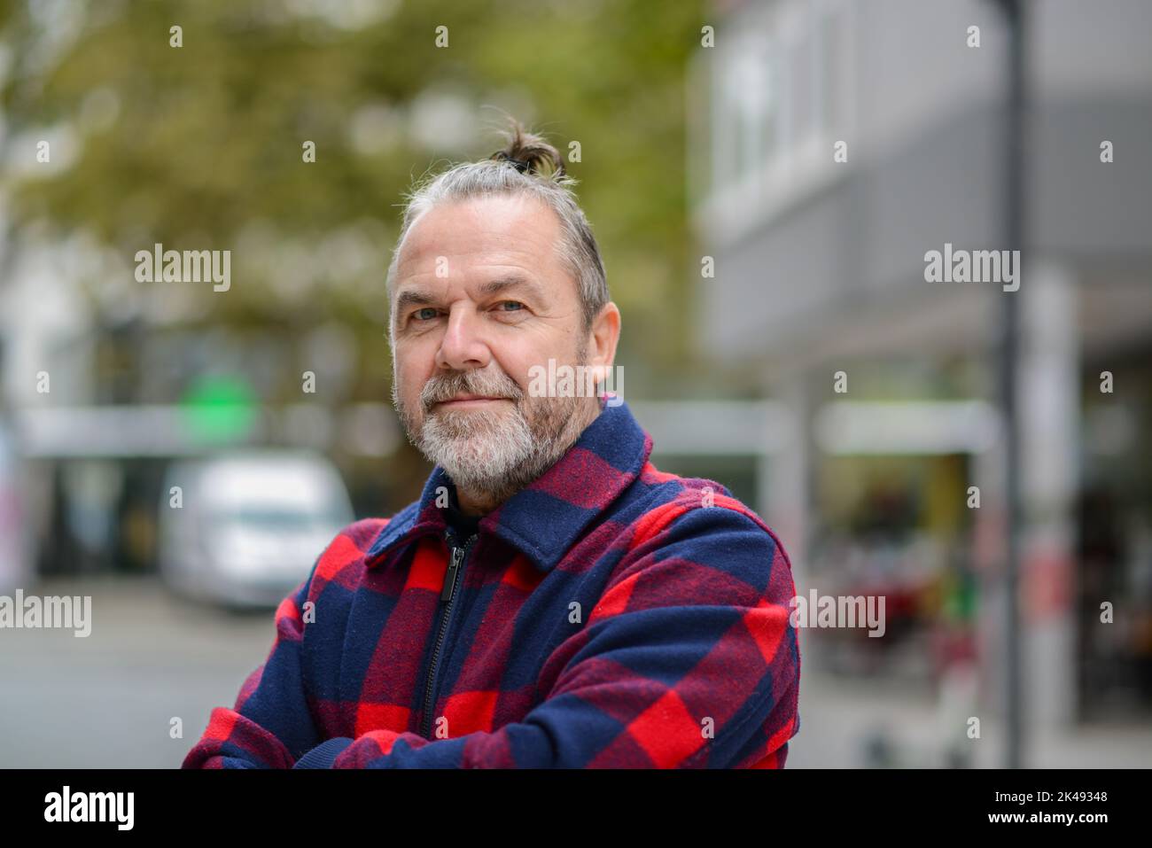 Uomo di mezza età con una mordente pistola in una giacca rossa e blu nello stile di un lumberjack si alza con le braccia incrociate in una strada commerciale Foto Stock