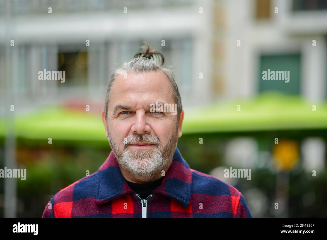 Uomo di mezza età con un sudicio mordente in una giacca in stile lumberjack rosso e blu si trova in una strada commerciale con un sorriso morbido Foto Stock