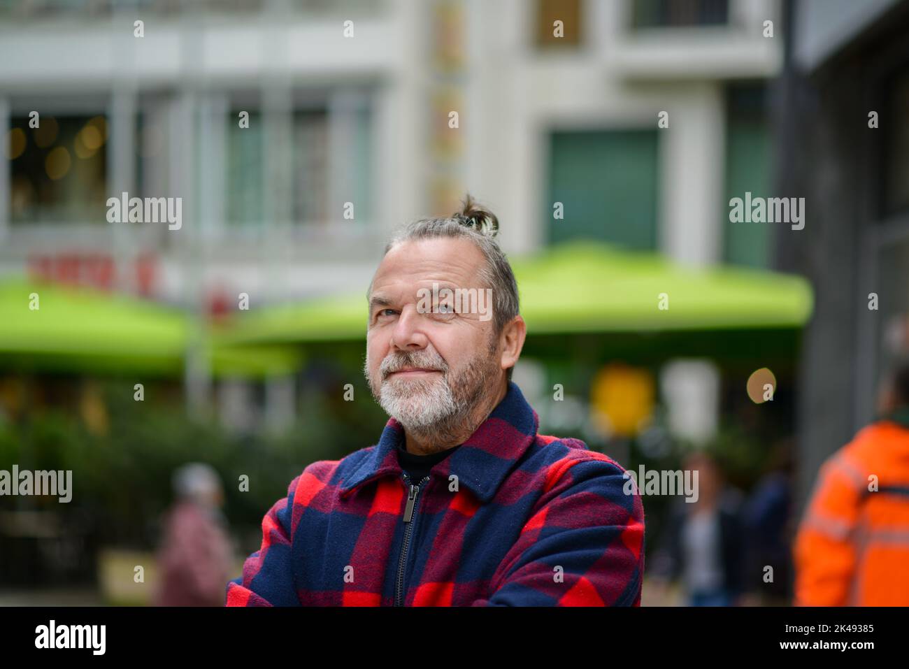 Uomo di mezza età con un sudicio mordente in una giacca rossa e blu nello stile di un lumberjack si alza con le braccia incrociate in una strada commerciale Foto Stock