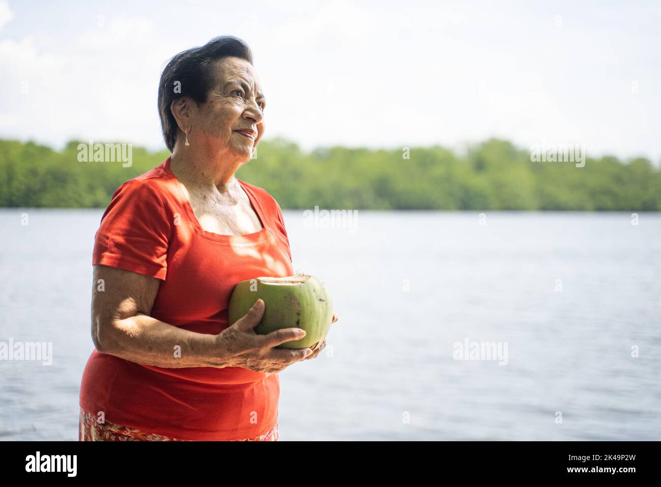 Donna anziana latina o ispanica che tiene una noce di cocco vicino a un lago, fiume o estuario. Alberi sullo sfondo. Foto Stock