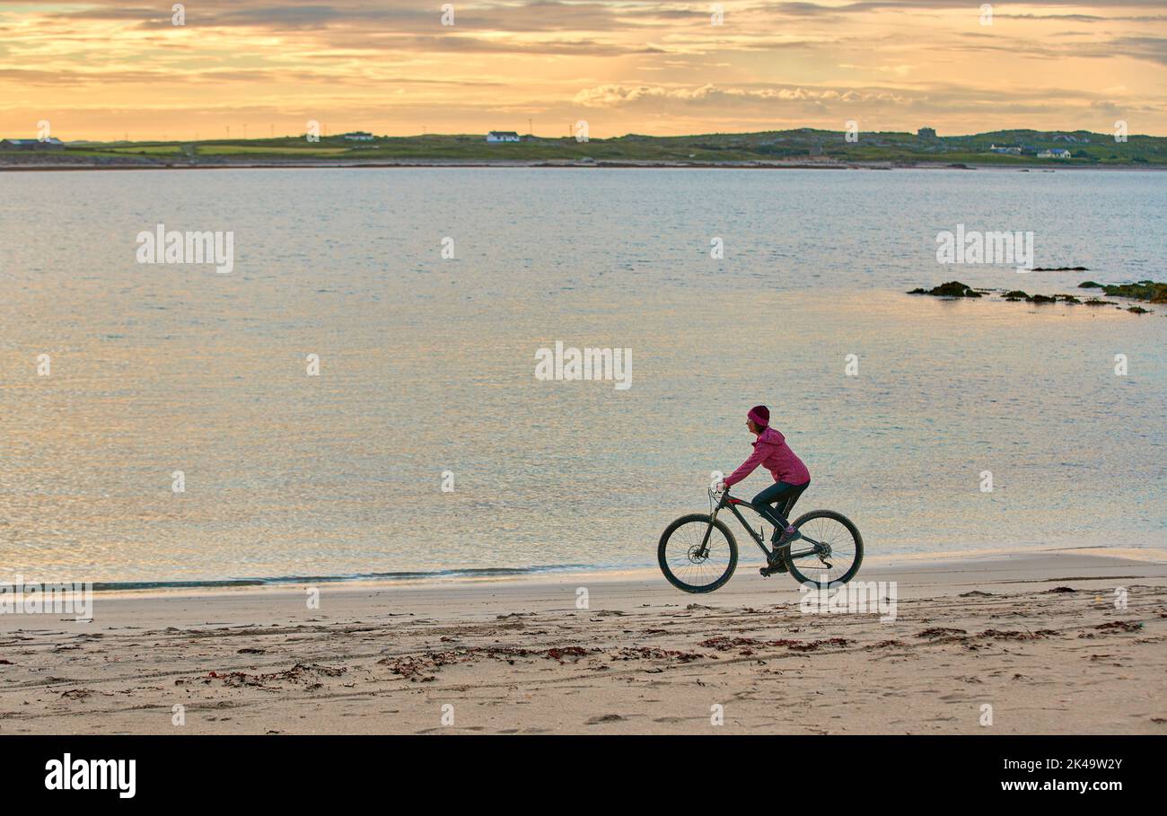 Bella donna anziana in mountain bike, in bicicletta al tramonto sulla spiaggia di sabbia dorata di Sillerna, Grallagh, County Galway, nella parte occidentale della Repubblica Foto Stock