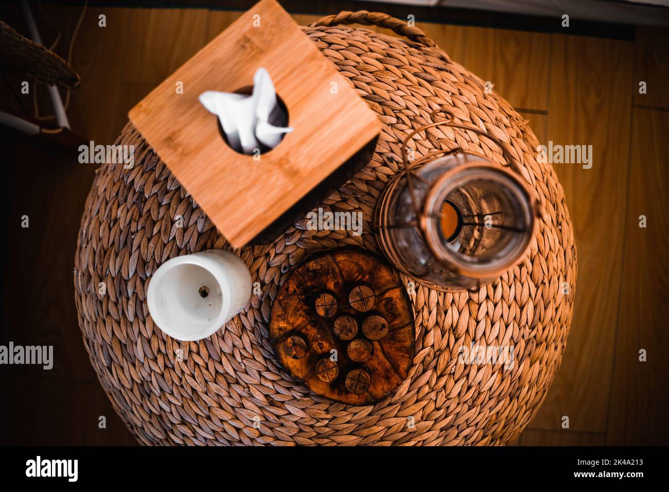 comodino rotondo in vimini su un pavimento in legno con una scatola di tessuti, una candela e un lampadario di vetro Foto Stock