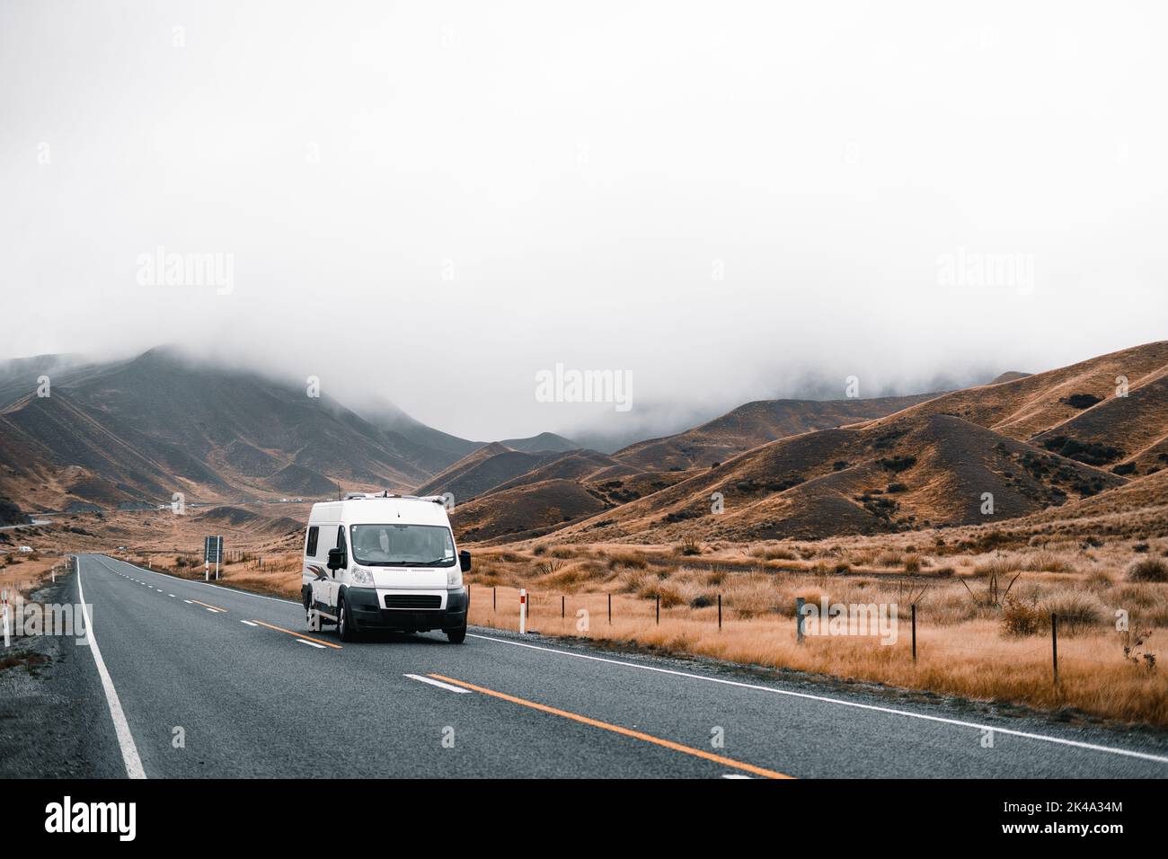 furgone in movimento solitario che guida lungo una lunga e stretta strada solitaria senza traffico tra montagne e campagna con poca vegetazione in una giornata di nebbia con Foto Stock