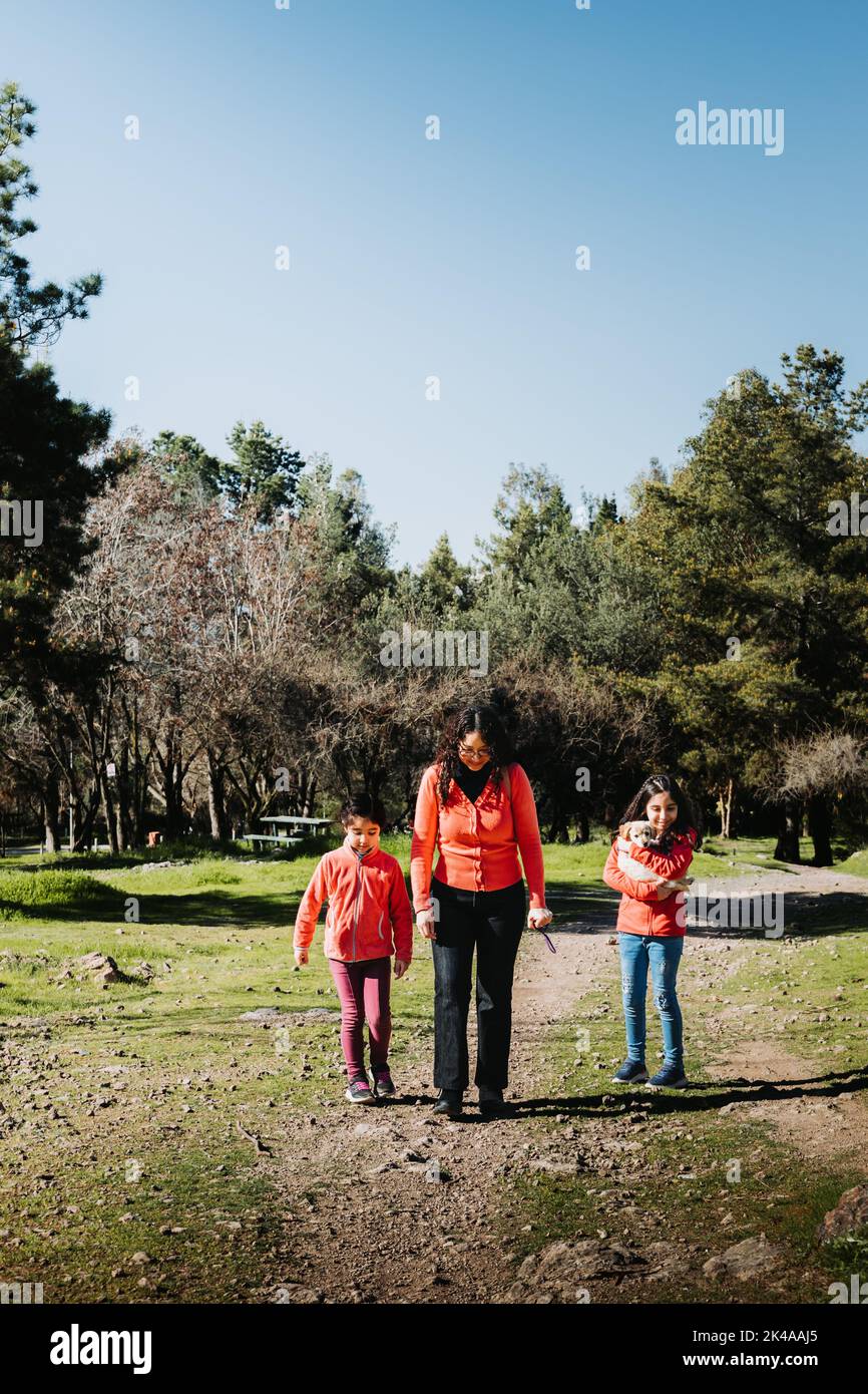 Giovane donna con le sue due figlie che camminano sull'erba e giocano con un cucciolo di recupero d'oro nel parco Foto Stock