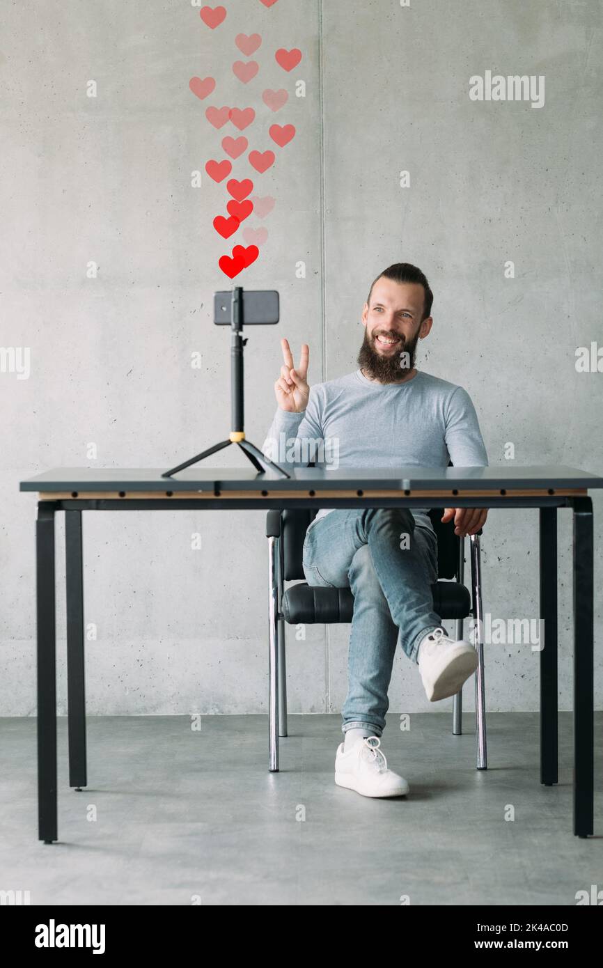 Influencer di successo. Rete come. Ammirazione degli spettatori. Sicuro sorridente vlogger maschile registrazione di contenuti video su smartphone che mostrano il gesto di pace V. Foto Stock
