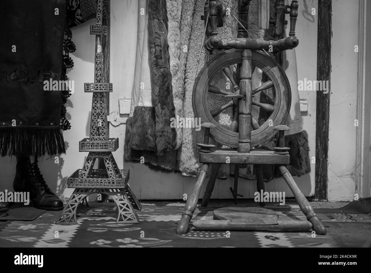 La vista in scala di grigi di una ruota rotante in legno, statuette, cappotti e stivali da donna sul tappeto di una stanza Foto Stock