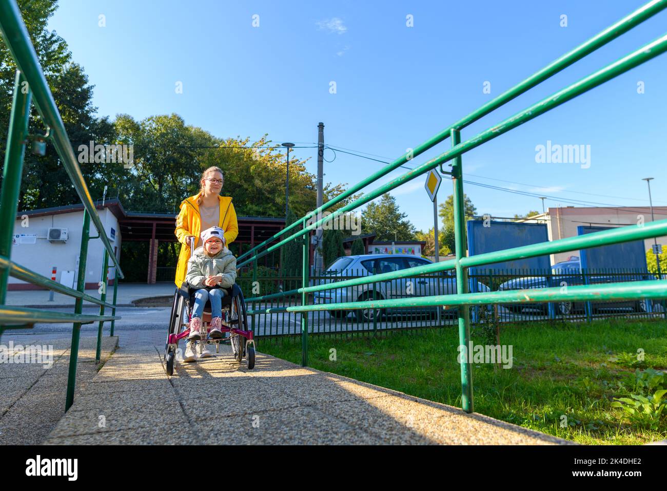 Giovane madre che spinge una sedia a rotelle con la figlia, ragazza che vive con paralisi cerebrale, sulla loro strada per la terapia. Accesso senza barriere architettoniche, sedia a rotelle o a rotelle Foto Stock