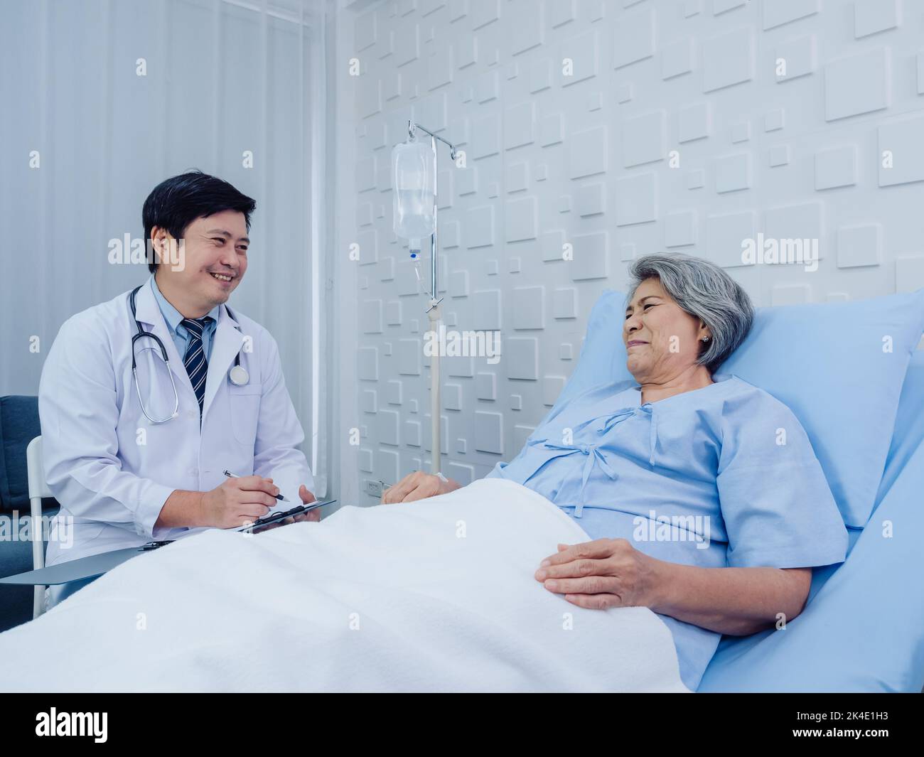 Sorridente medico asiatico in tuta bianca prendere appunti mentre si discute felice asiatico anziano, donna anziana paziente che sdraiato sul letto con la terapia salina soluti Foto Stock