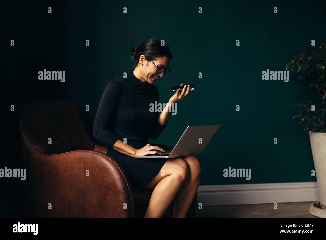 Felice donna asiatica che risponde a una telefonata e lavora sul computer portatile. Giovane donna seduta in poltrona con laptop e telefono cellulare. Foto Stock